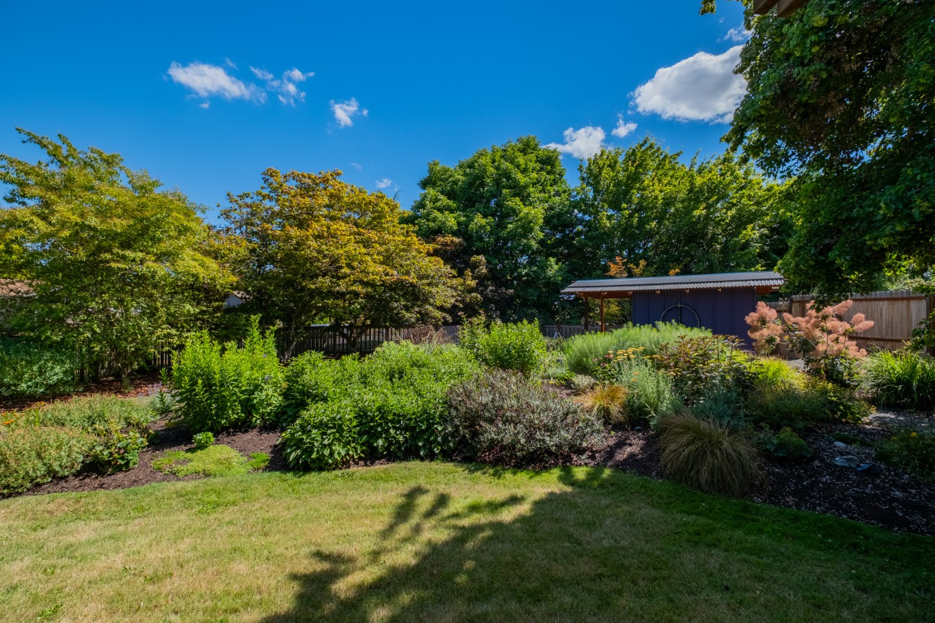 Fenced backyard with mature planting beds, a lawn, and a detached garden studio