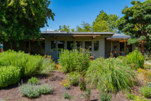 Back of the single-story furnished Corvallis home seen across a planted perennial garden, with a wall of windows and a covered patio with Adirondack chairs