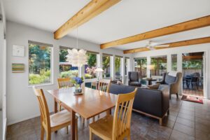 Sunlit dining area with a wood table under a beamed ceiling, open to the living room and garden windows